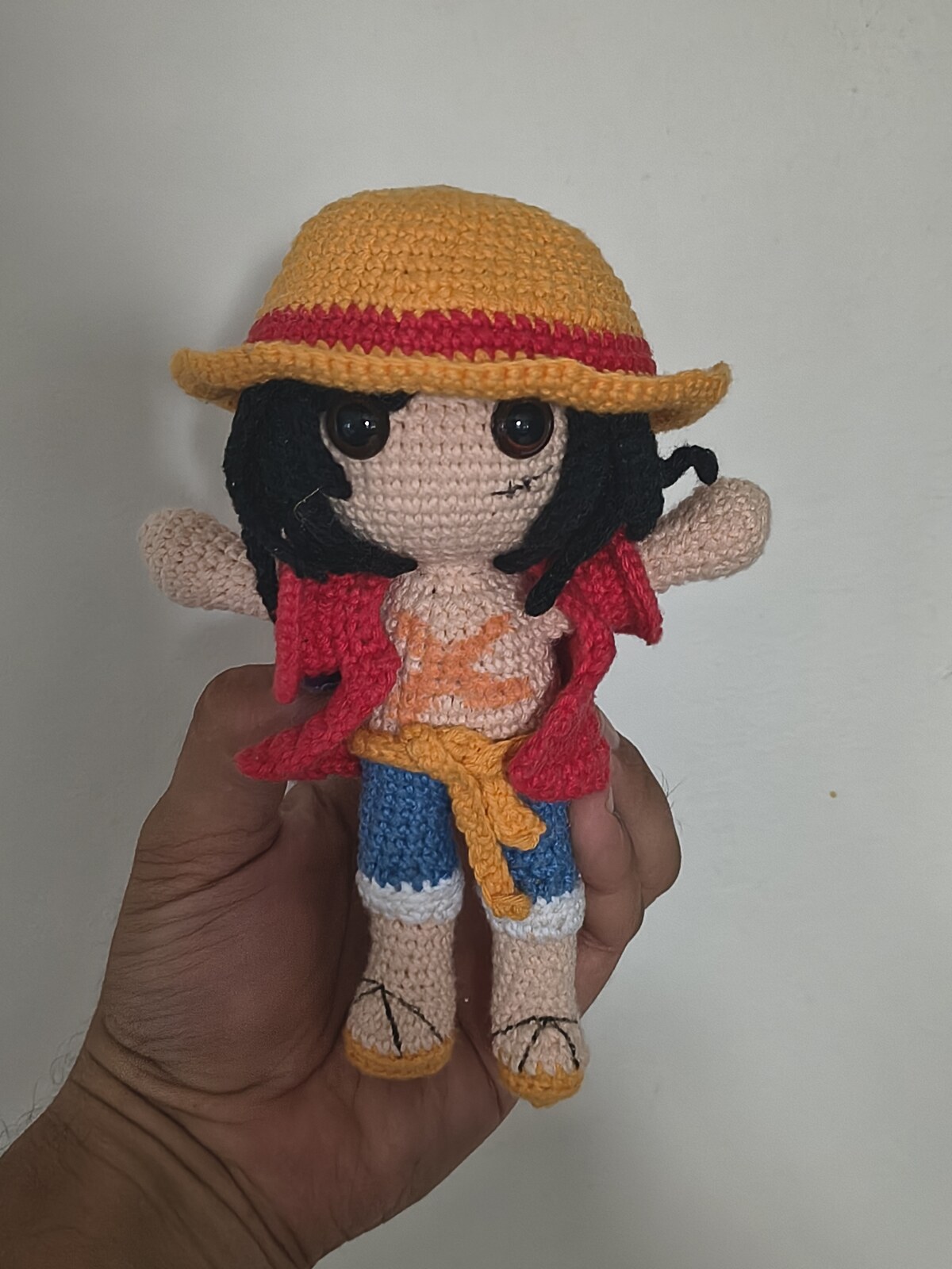 L'amigurumi de Monkey D. Luffy terminé, tenu dans une main, avec son chapeau de paille, son gilet rouge, sa ceinture jaune et son short bleu