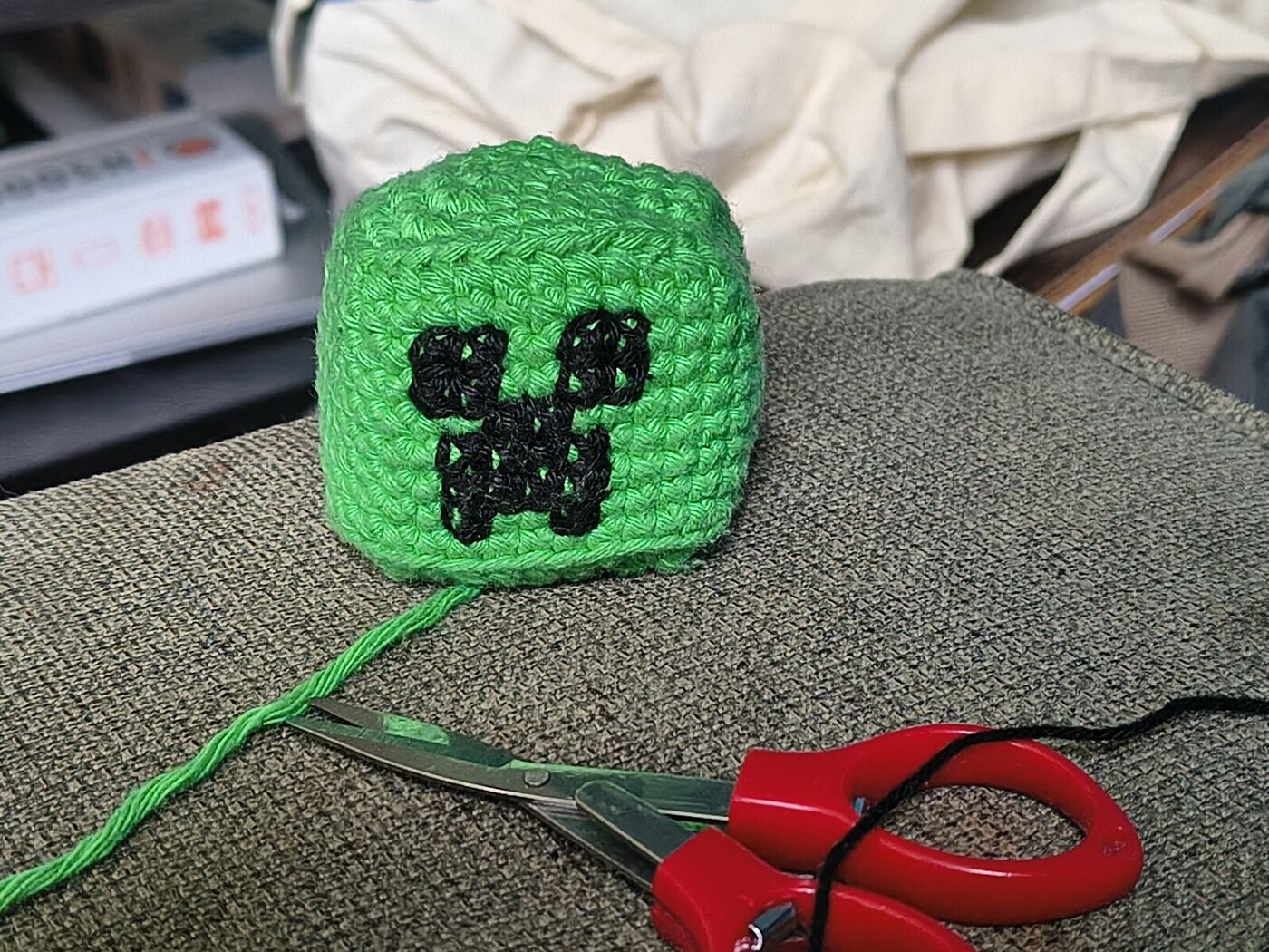 Travail en cours : la tête cubique verte au crochet avec le visage du Creeper brodé en laine noire, à côté de ciseaux rouges, du fil vert et d'une aiguille enfilée de laine noire